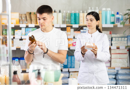 Guy chooses cough syrup in modern pharmacy. Female pharmacist helps with choosing medicine Guy chooses cough syrup in modern pharmacy. Female pharmacist helps with choosing medicine 134471311