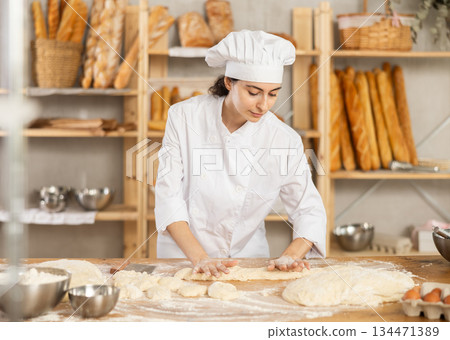 Young woman works as baker, mold baguette from dough, creates long loafs from dough. Young woman works as baker, mold baguette from dough, creates long loafs from dough. 134471389