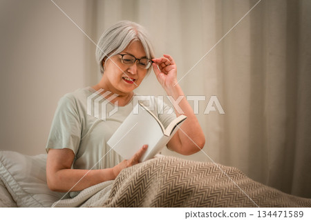 A gray-haired senior woman reading in her bedroom before going to sleep. 134471589