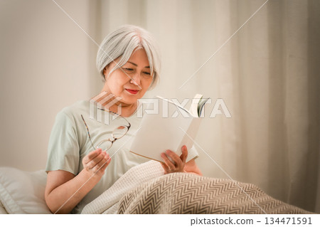 一位頭髮花白的老婦人在睡前於臥室裡閱讀。 134471591