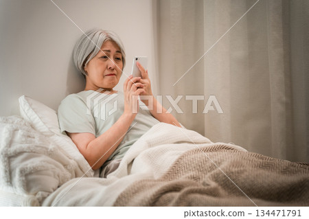 A gray-haired senior woman looking at her smartphone before going to sleep in her bedroom (troubled, worried, anxious, worried) 134471791