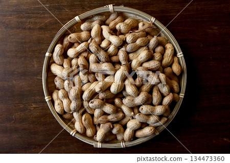 A pile of shelled peanuts in a bamboo basket with a table in the background, viewed from directly above 134473360