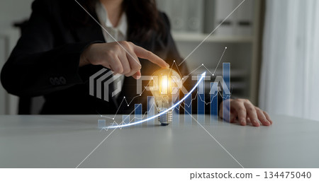 Businesswoman in Suit Pointing at Light Bulb with Rising Graphs, Illustrating Innovation and Growth in Corporate Strategy and Decision-Making Process Scalp 134475040