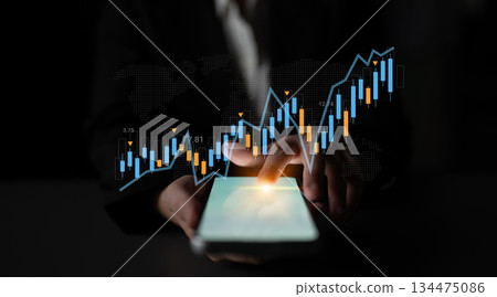 Business professional using smartphone to analyze financial data with rising graph, stock market trends, and digital analytics in dark background Gantry 134475086