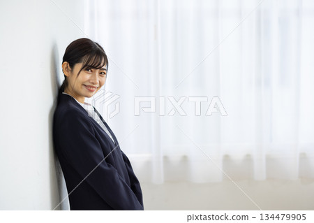 一位身著西裝的年輕日本女商務人士倚靠在白牆上，面帶微笑。 134479905