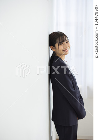 A young Japanese female business person in a suit leaning against a white wall and smiling A young Japanese female business person in a suit leaning against a white wall and smiling 134479907