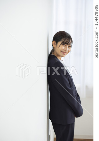 一位身著西裝的年輕日本女商務人士倚靠在白牆上，面帶微笑。 134479908