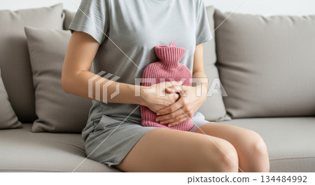 Woman sitting on a sofa, holding a hot water bottle to her abdomen, expressing discomfort from menstruation pain and seeking relief at home 134484992
