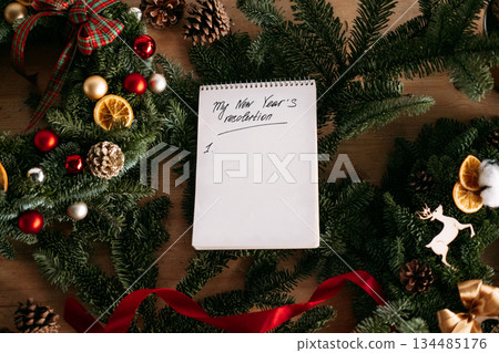 Blank New Years resolution list on a notepad surrounded by festive evergreen decorations. Goal setting, personal growth, mental health reflection, seasonal motivation 134485176