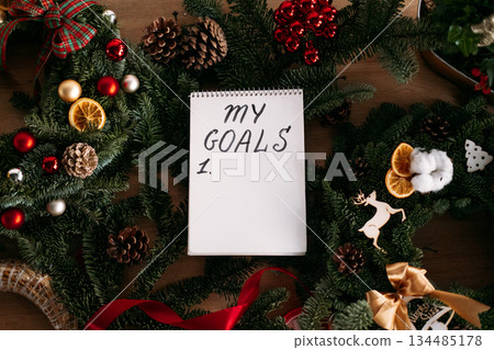 Title: Spiral notebook displaying MY GOALS list on wooden table among festive evergreen branches and Christmas decorations. New Year goals, personal planning, holiday reflection, self-improvement Title: Spiral notebook displaying MY GOALS list on wooden table among festive evergreen branches and Christmas decorations. New Year goals, personal planning, holiday reflection, self-improvement 134485178