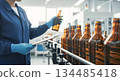 Worker in blue lab coat inspecting amber glass bottles on production line, ensuring quality control in beverage manufacturing process 134485418