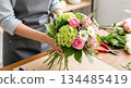 Florist arranging a vibrant bouquet of roses, hydrangeas, and daisies on a wooden table, showcasing floral artistry and creativity in a bright workspace 134485419