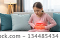 Young girl in pink pajamas is sitting on a couch, holding her stomach in discomfort, with soft pillows and warm lighting creating a cozy atmosphere 134485434
