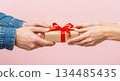 Hands exchanging a beautifully wrapped gift with a red ribbon against a soft pink background, celebrating love and connection on Valentine's Day 134485435