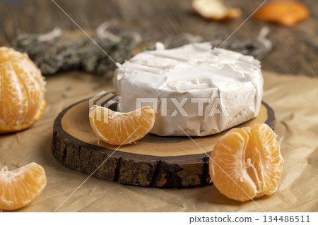 fresh white cheese with white mold lies on a wooden board on crumpled yellow paper, soft round cheese and juicy sweet orange tangerines fresh white cheese with white mold lies on a wooden board on crumpled yellow paper, soft round cheese and juicy sweet orange tangerines 134486511