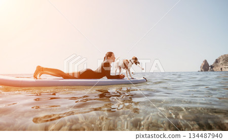 Woman Dog Paddleboard Sunset - Woman and dog paddleboarding together on a calm ocean at sunset. 134487940