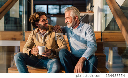 Two friends share laughter and warm drinks on a wooden staircase in a cozy home setting during daylight hours 134488554