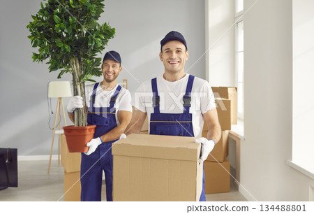 Mover workers in uniform holding a cardboard box and green plant looking at camera 134488801