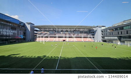 A vibrant stadium with a soccer field and grandstand sits beside the Festive Arts Theatre, blending athletic energy and cultural architecture in a dynamic 134489014