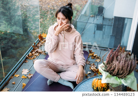 Woman practices Nadi Shodhana pranayama while sitting on yoga mat on balcony with autumn leaves. Mindfulness, outdoor wellness, mental health. 134489123
