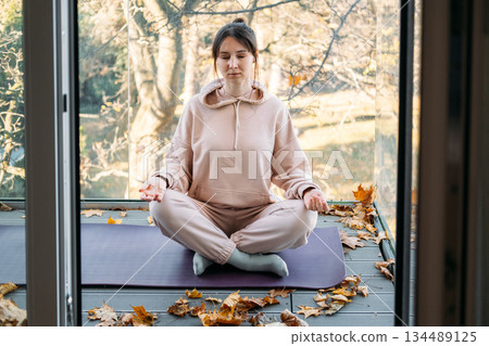 Woman sits with eyes closed in meditative pose on balcony during autumn. Peaceful solitude, intentional living, mental space, digital detox. 134489125