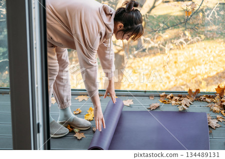 Woman stretches side on yoga mat among autumn leaves on balcony. Balcony yoga, small space fitness, terrace workout. Woman stretches side on yoga mat among autumn leaves on balcony. Balcony yoga, small space fitness, terrace workout. 134489131