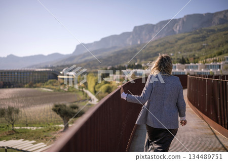 Woman walking mountains railing overlooking vast scenic landscape and modern resort Woman walking mountains railing overlooking vast scenic landscape and modern resort 134489751