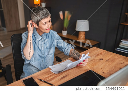 Focused mature businesswoman reviewing documents at home office desk Focused mature businesswoman reviewing documents at home office desk 134491011