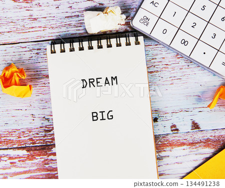 A notepad shows the text DREAM BIG. A calculator rests nearby, along with crumpled paper, implying planning and aspiration on a distressed, colorful wooden surface A notepad shows the text DREAM BIG. A calculator rests nearby, along with crumpled paper, implying planning and aspiration on a distressed, colorful wooden surface 134491238