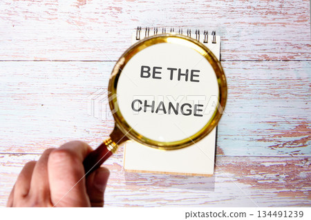 Someone holds a magnifying glass over a notebook. The notepad displays the motivational words BE THE CHANGE. The backdrop features an aged, wooden surface, suggesting age and wisdom Someone holds a magnifying glass over a notebook. The notepad displays the motivational words BE THE CHANGE. The backdrop features an aged, wooden surface, suggesting age and wisdom 134491239