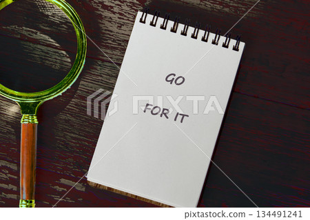 A notepad displaying GO FOR IT rests near a golden magnifying glass on a weathered wood surface. This scene inspires motivation and exploration of possibilities A notepad displaying GO FOR IT rests near a golden magnifying glass on a weathered wood surface. This scene inspires motivation and exploration of possibilities 134491241