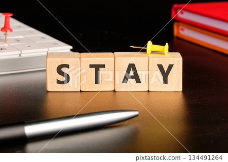 Wooden blocks spell out STAY on a desk. A pen and computer are nearby, suggesting a work environment. Push pins add a touch of visual flair 134491264