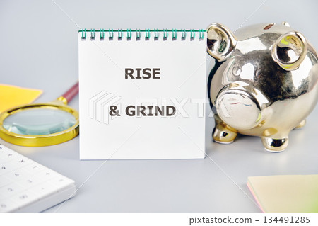 A spiral notepad displaying the words RISE GRIND stands next to a golden Piggy Bank. A magnifying glass, calculator and sticky notes complete the scholastic scene A spiral notepad displaying the words RISE GRIND stands next to a golden Piggy Bank. A magnifying glass, calculator and sticky notes complete the scholastic scene 134491285