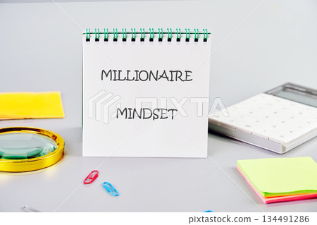 A notepad displays Millionaire Mindset, surrounded by a calculator, sticky notes, paperclips and magnifying glass, symbolizing strategic planning on a grey surface 134491286