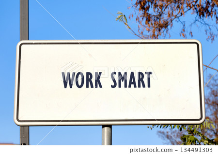 A white sign displays the words Work Smart in dark blue lettering. The sign is mounted on a pole, set against a radiant blue sky with trees in the background A white sign displays the words Work Smart in dark blue lettering. The sign is mounted on a pole, set against a radiant blue sky with trees in the background 134491303