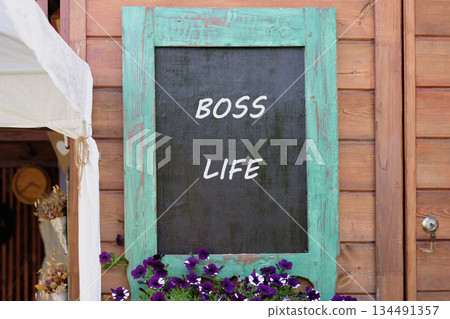 A vintage blackboard displays the words Boss Life in white. Purple flowers flourish below, and the rustic wooden wall adds warmth. It evokes a sense of carefree style 134491357