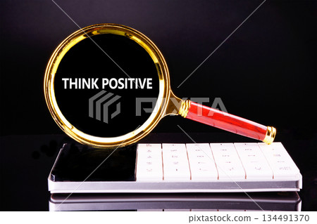 A golden magnifying glass with THINK POSITIVE displayed in the lens rests on a white and gray calculator against a dark reflective backdrop, creating a striking visual 134491370