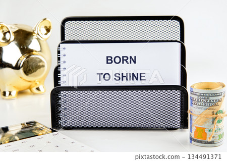 On a white table, a notebook with Born To Shine sits next to a roll of United States currency, a golden piggy bank, and a calculator, symbolizing financial aspirations 134491371