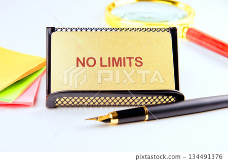 A golden card that reads NO LIMITS sits in a black holder. A Fountain Pen, sticky notes, and a magnifying glass complement the atmosphere of endless possibilities on a desk 134491376