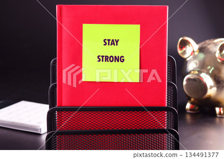 A red book sits in a black metal organizer, displaying a yellow sticky note with the phrase STAY STRONG. A golden piggy bank sits on the desk nearby A red book sits in a black metal organizer, displaying a yellow sticky note with the phrase STAY STRONG. A golden piggy bank sits on the desk nearby 134491377