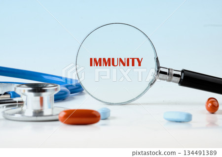 Magnifying glass focuses on the word Immunity, shown in red text. Stethoscope and pills surround the glass, against a light blue backdrop representing healthcare research Magnifying glass focuses on the word Immunity, shown in red text. Stethoscope and pills surround the glass, against a light blue backdrop representing healthcare research 134491389