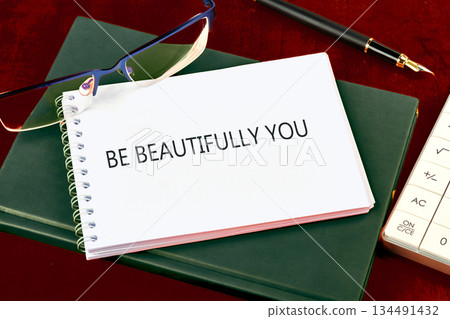 A spiral notebook displaying the phrase BE BEAUTIFULLY YOU sits atop a green journal, accompanied by glasses, a fountain pen, and a calculator on a dark wooden desk 134491432