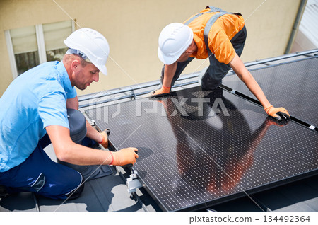 Workers building solar panel system on rooftop of house. Two men installers in helmets installing photovoltaic solar module outdoors. Alternative, green and renewable energy generation concept. Workers building solar panel system on rooftop of house. Two men installers in helmets installing photovoltaic solar module outdoors. Alternative, green and renewable energy generation concept. 134492364