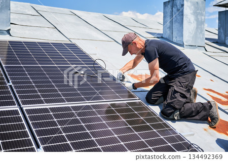 Worker building solar panel system on metal rooftop of house. Man engineer installing photovoltaic solar module outdoors. Alternative, green and renewable energy generation concept. 134492369