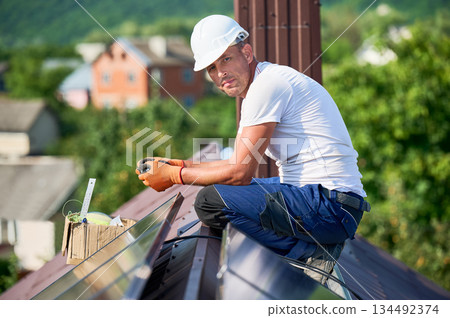 Worker building solar panel system on metal rooftop of house. Portrait of engineer installing photovoltaic solar module outdoors. Alternative, green and renewable energy generation concept. Worker building solar panel system on metal rooftop of house. Portrait of engineer installing photovoltaic solar module outdoors. Alternative, green and renewable energy generation concept. 134492374