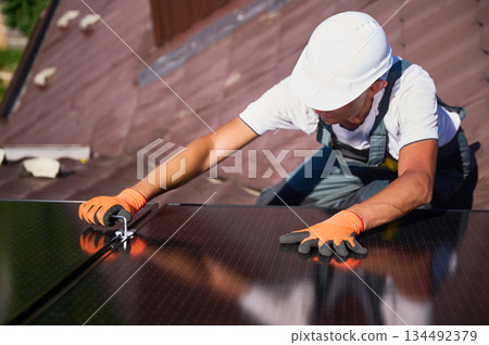Worker building photovoltaic solar panel system on rooftop of house. Close up of man engineer in helmets and gloves installing solar module with help of hex key outdoors. Renewable energy. 134492379
