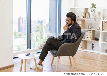 An Indian businessman is deep in thought while working on his laptop in a modern office setting An Indian businessman is deep in thought while working on his laptop in a modern office setting 134494126