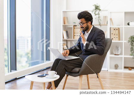 An Indian businessman gestures while participating in a video conference on his laptop in a modern office setting 134494147