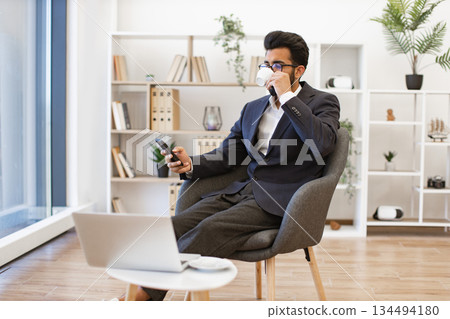 An Arab businessman is seen relaxing in a modern office setting, holding a phone and drinking coffee 134494180