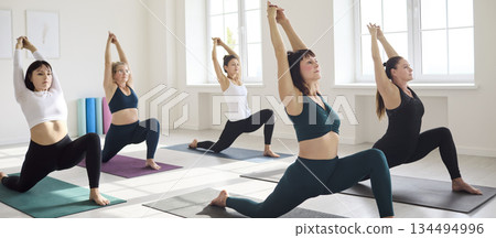 Women in yoga class hold lunge asana with arms raised, stretching together on mats Women in yoga class hold lunge asana with arms raised, stretching together on mats 134494996
